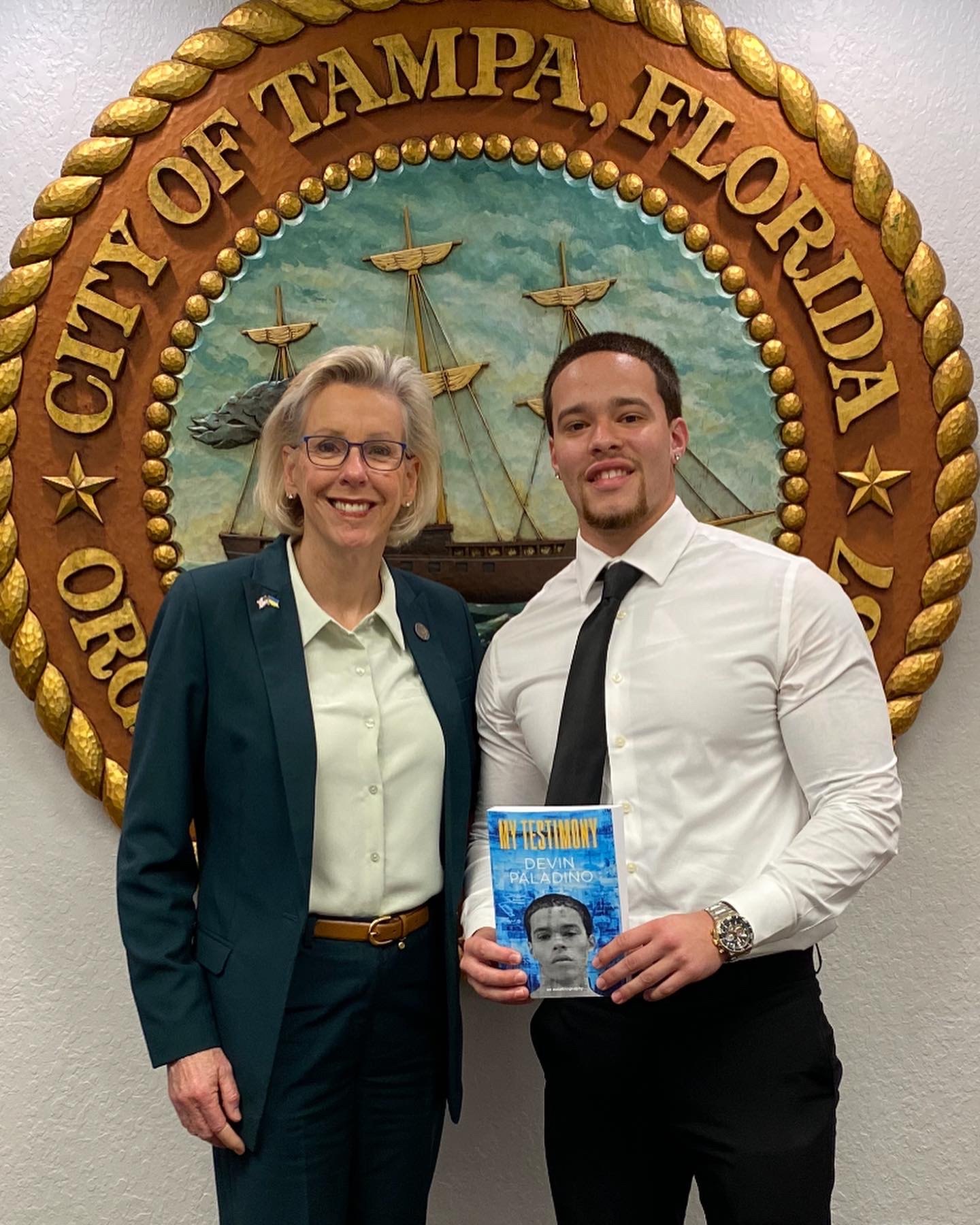 Devin Paladino holding a copy of My Testimony beside a city official in front of the City of Tampa seal