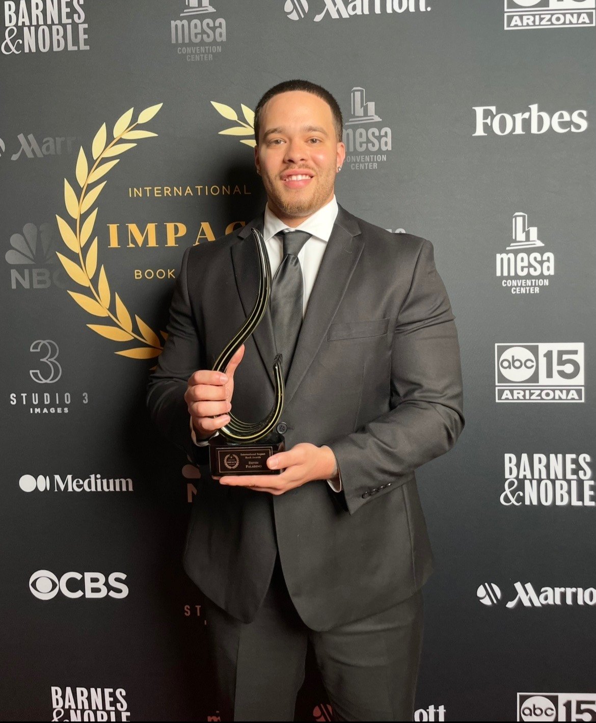 Devin Paladino in a suit, holding a trophy on the step-and-repeat at the International Impact Book Awards