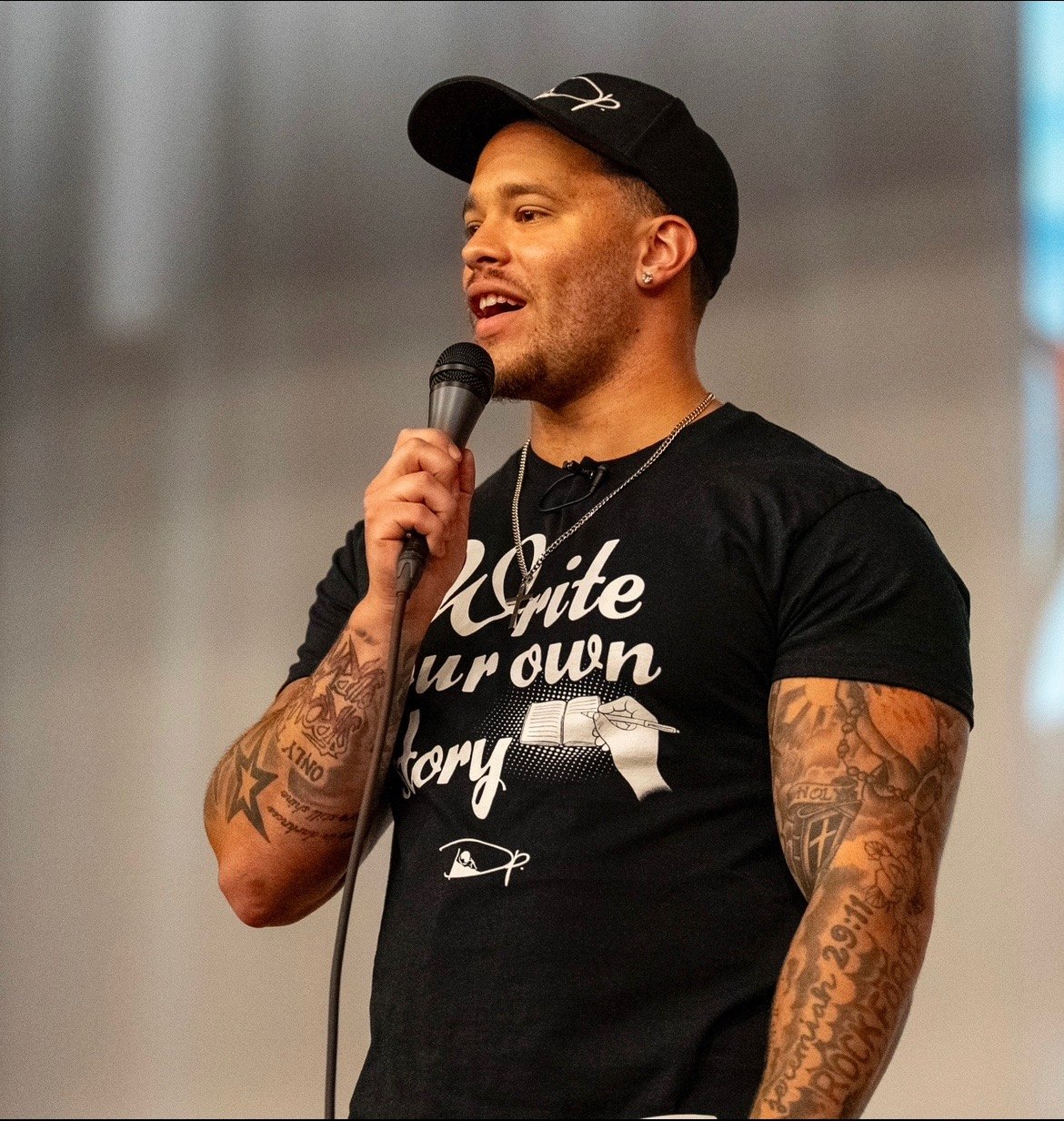 Close-up of Devin Paladino speaking into a handheld microphone, wearing a “Write Your Own Story” T-shirt