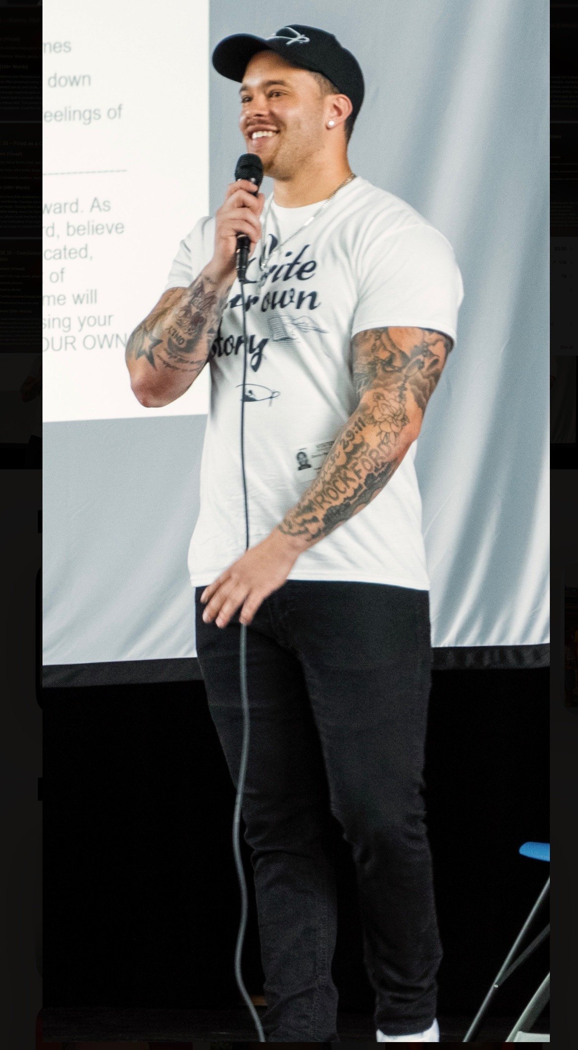 Devin Paladino on stage with a handheld microphone, a slide from his talk projected behind him