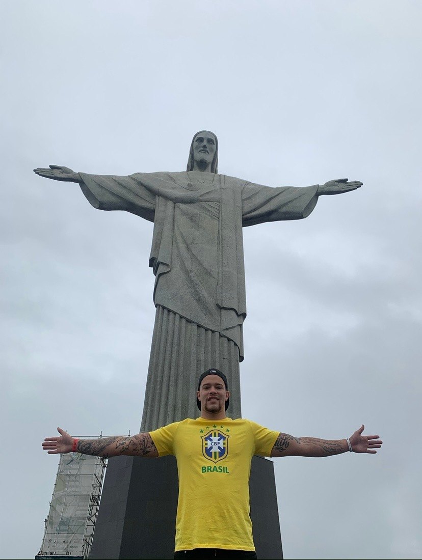 Devin Paladino below Christ the Redeemer in Rio de Janeiro, arms outstretched, in a Brazil shirt