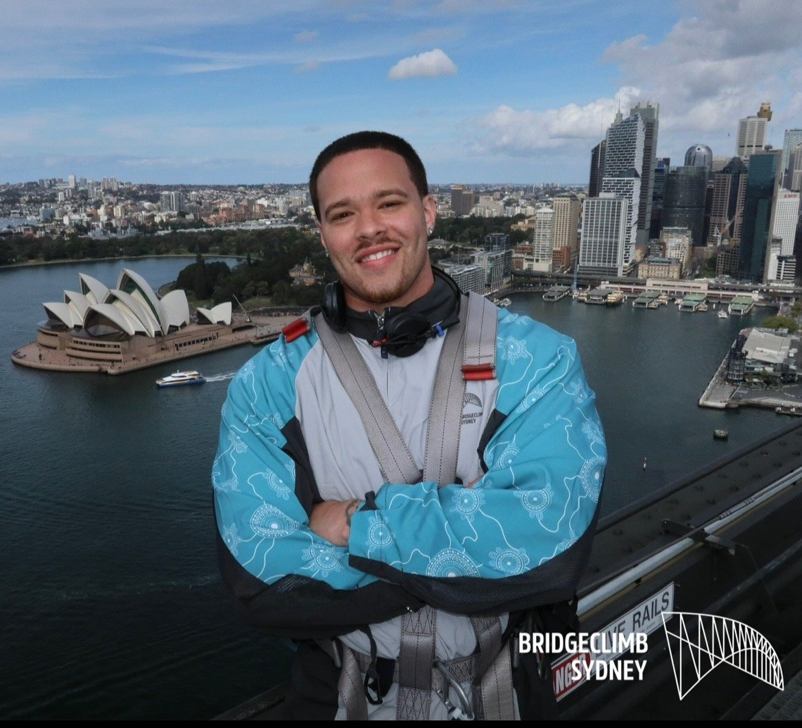 Devin Paladino in a BridgeClimb harness above Sydney Harbour, the Opera House behind him